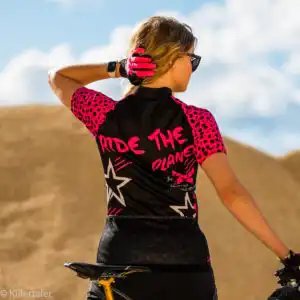 Rückansicht des Killertaler Damen Radtrikots kurzarm Travel in Schwarz mit pinkem Ride the Planet Schriftzug und dreiteiliger Rückentasche