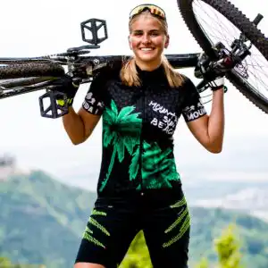 Sportliche Frau trägt ein Mountainbike auf den Schultern vor der Burg Hohenzollern, bekleidet mit dem grünen Damen Radtrikot Palm Tree und der grünen Baggy Bike Shorts Epic Ride