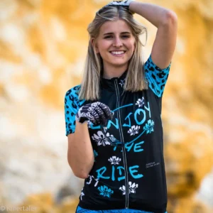 Eine lachende blonde Frau vor einem goldgelben Felsenhintergrund trägt das türkisch-schwarze Damen Fahrradshirt Enjoy the Ride