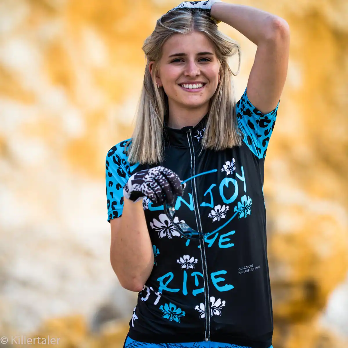 Eine lachende blonde Frau vor einem goldgelben Felsenhintergrund trägt das türkisch-schwarze Damen Fahrradshirt Enjoy the Ride