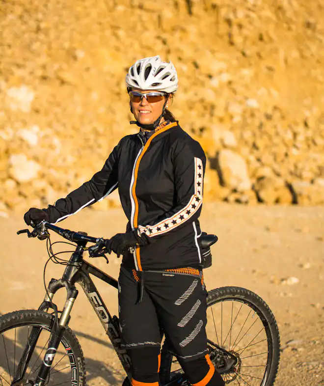 Damen Fahrrad Windjacke Mavora in Schwarz-Orange mit Kapuze und elastischem Stretch neben einem Mountainbike in einer Felslandschaft. Perfekt angepasst dank Taillen- und Armlängen-Maßanfertigung.